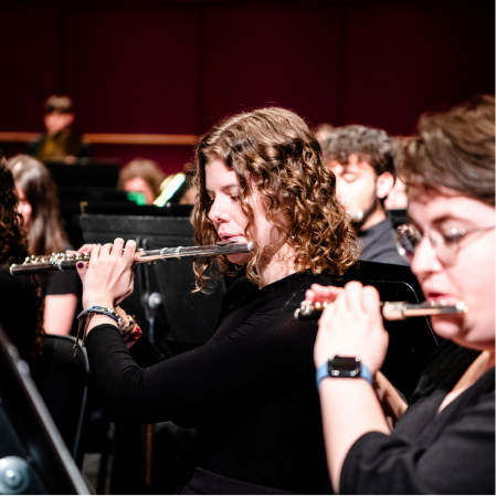 A person playing the flute as part of an orchestra, with other musicians and music stands visible in the background