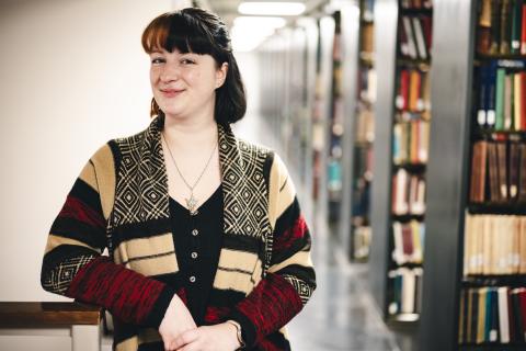 Olivia Mathers stands in the stacks and poses for a photo. She wears a stripped cardigan.