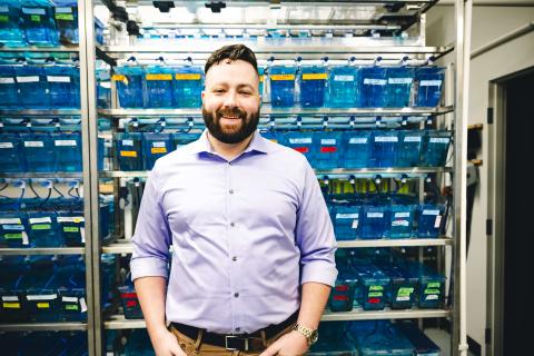 Alex Seaver stands in front of an array of little fish tanks wearing a purple button up shirt.