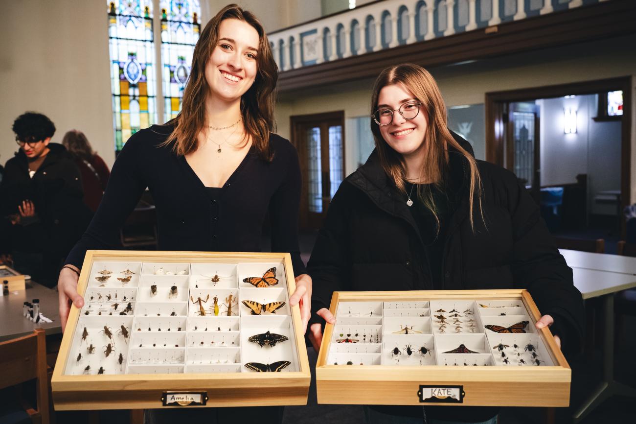 Two female students stand in a classroom holding their insect frames
