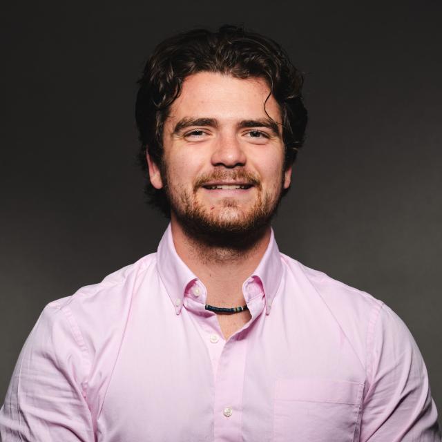 Aidan McKee wearing a pink button down shirt smiling for his headshot in front of a black backdrop.