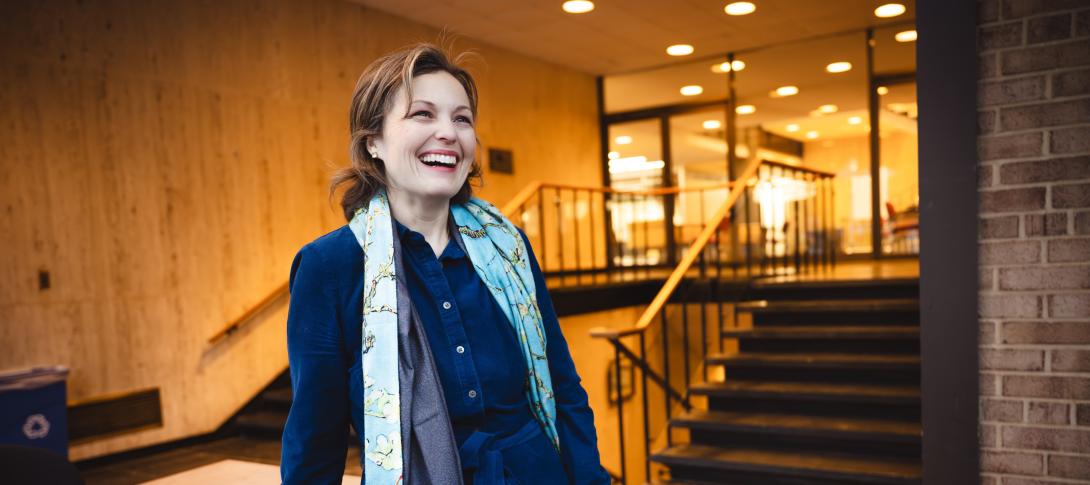 Liz Barrett stands in front of steps wearing a dark blue dress and looks off the the side laughing candidly.