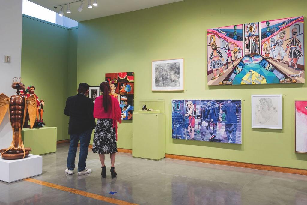 A man and a woman stand with their backs to the camera, viewing a diverse collection of colorful paintings and sculptures displayed on a lime green wall in a brightly lit art gallery.