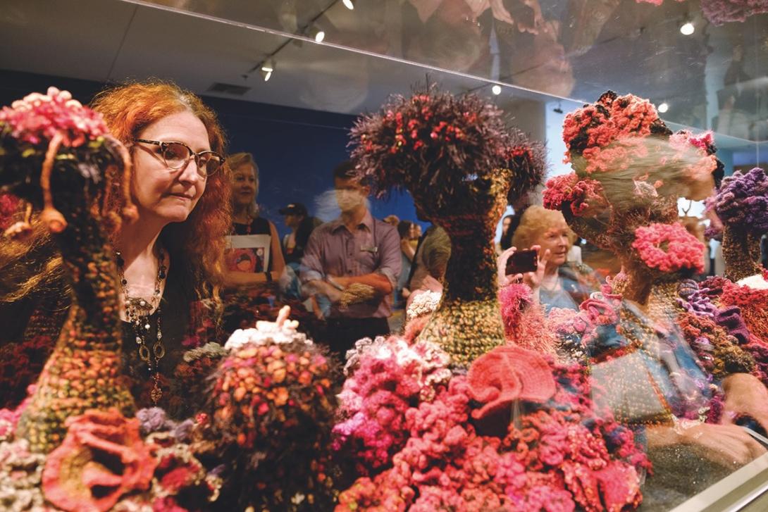 A woman examines a vibrant, crocheted coral reef installation inside a glass display case at an art museum.