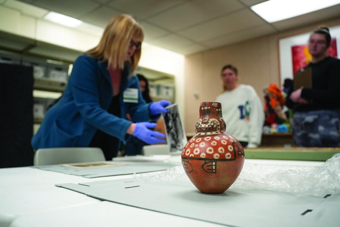  In an indoor classroom or lab setting, a patterned red and white ceramic vessel sits in the foreground on a white table. In the blurred background, a woman wearing blue gloves handles a document while students observe.