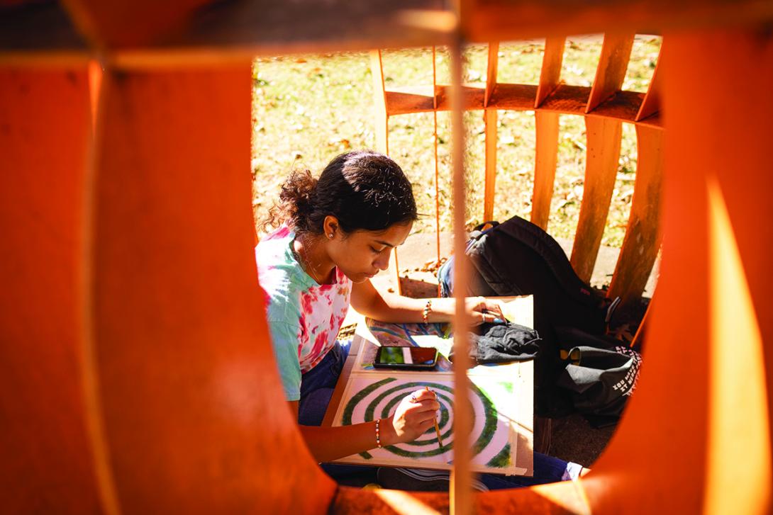 A student in a tie-dye shirt sits inside a large, orange outdoor sculpture, focused on painting a green spiral design on a canvas. The scene is captured through the curved openings of the structure, with sunlight illuminating the workspace.